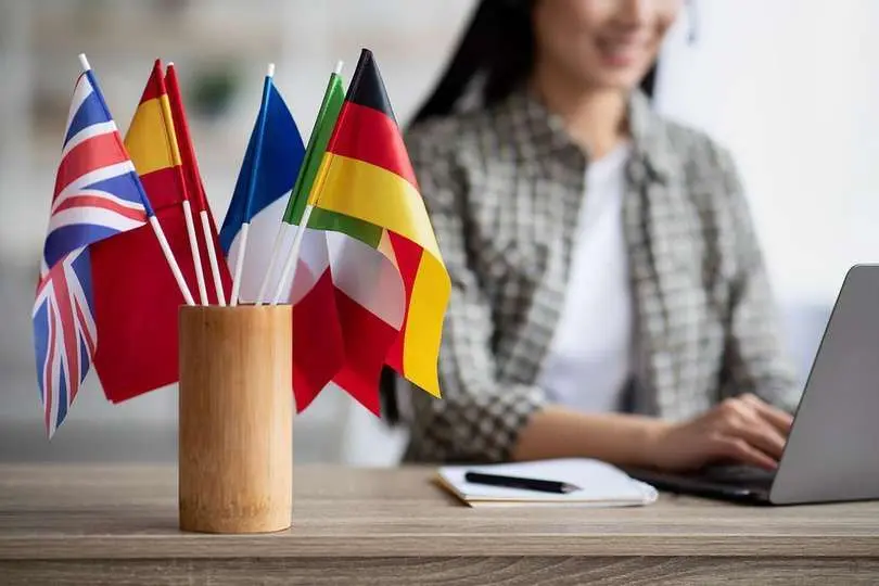 Cropped of asian young woman foreign language teacher sitting at workdesk and using laptop, selective focus on international flags. Foreign language online course, class, e-education concept